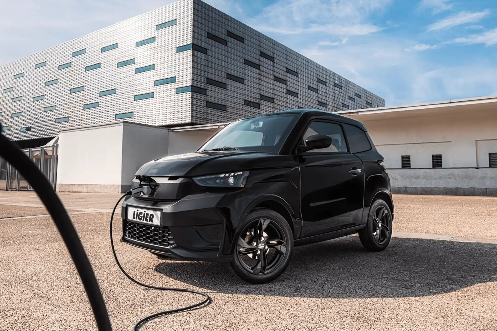 Ligier JS60 noire branchée à un câble de recharge devant un bâtiment moderne